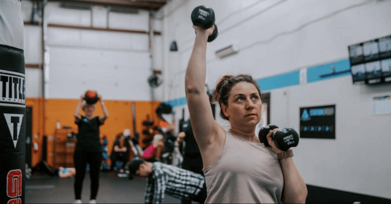 A woman pressing a dumbbell overhead during a HIIT circuit class at Applied Fitness Solutions in Ann Arbor, with other members working at step and floor stations behind her.